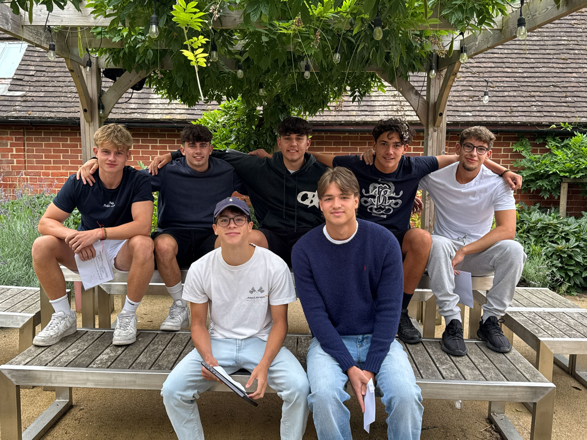 Seven gents pose together on wooden benches having received their A Level results