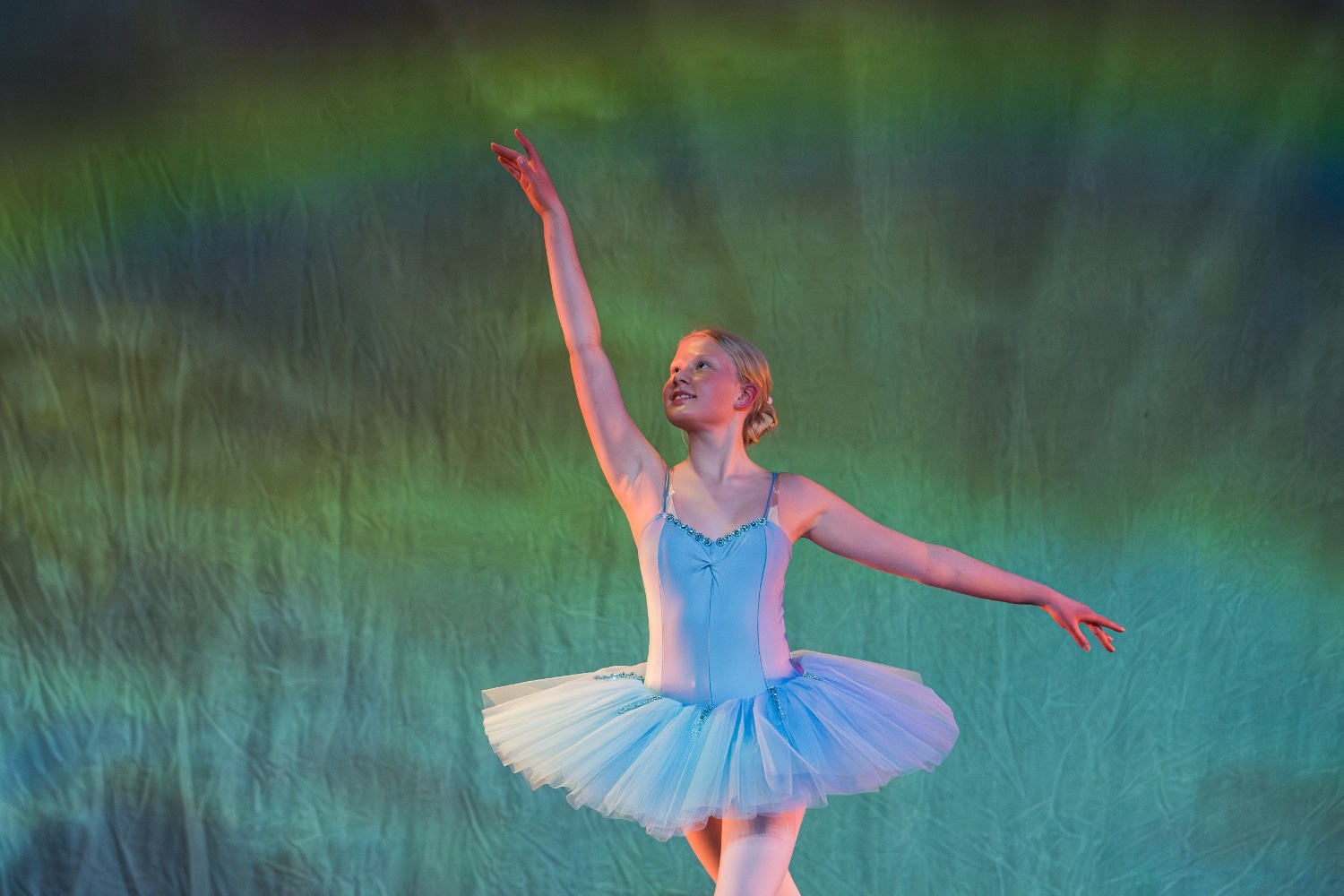 A girl ballet dancer in a blue tutu stands en pointe with one arm raised gracefully and the other extended to the side, performing on stage against a green and blue softly lit background.