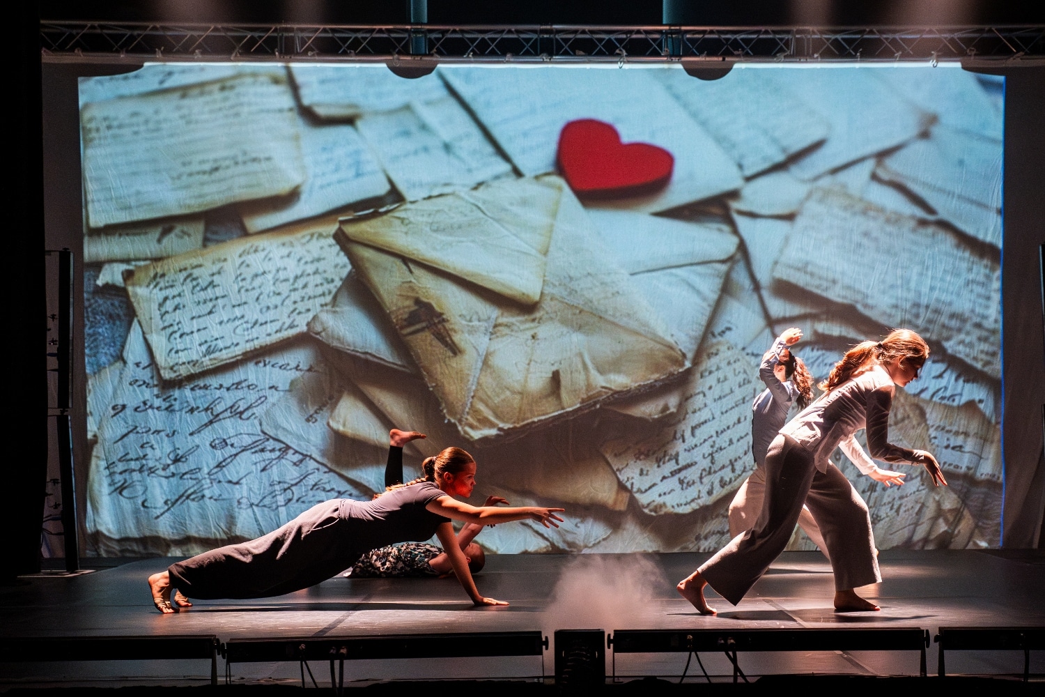 Three dancers perform expressively on stage in front of a large backdrop showing old handwritten letters with a red heart on top, creating an atmosphere of nostalgia and emotion.