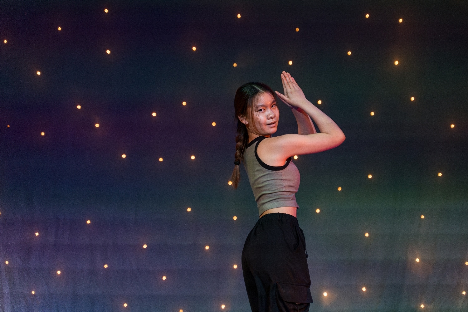 A young dancer poses with her hands together above her head, standing sideways in front of a backdrop decorated with small, glowing lights. She wears a grey tank top and black trousers.