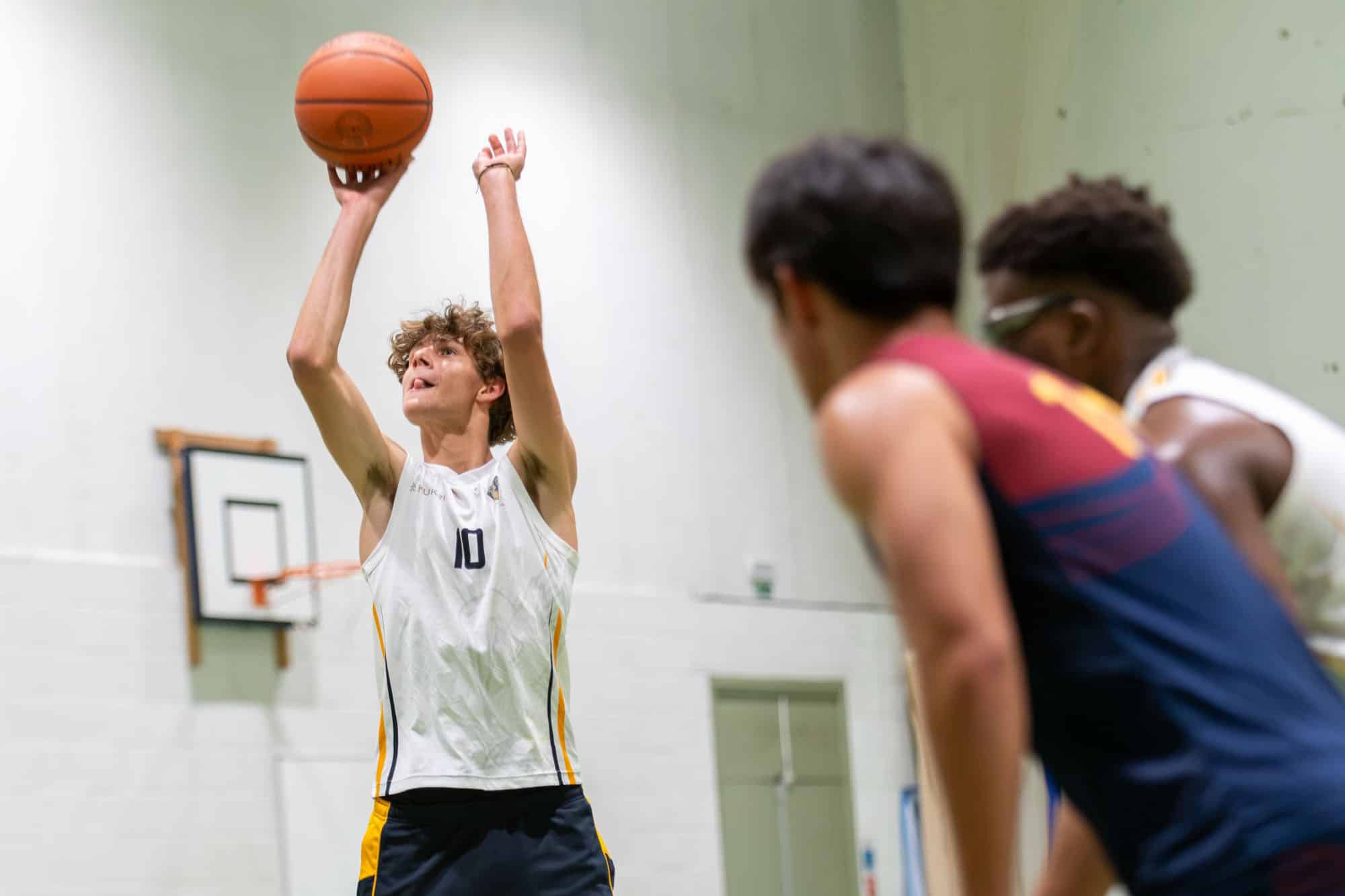 Two basketball players manoeuvring on an indoor court, with one player in a white jersey dribbling the ball while the other in a navy and maroon jersey defends.