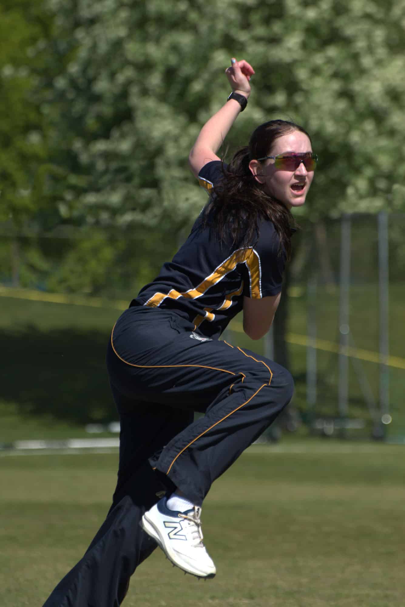 A cricket player in a navy and gold kit mid-delivery on a pitch.