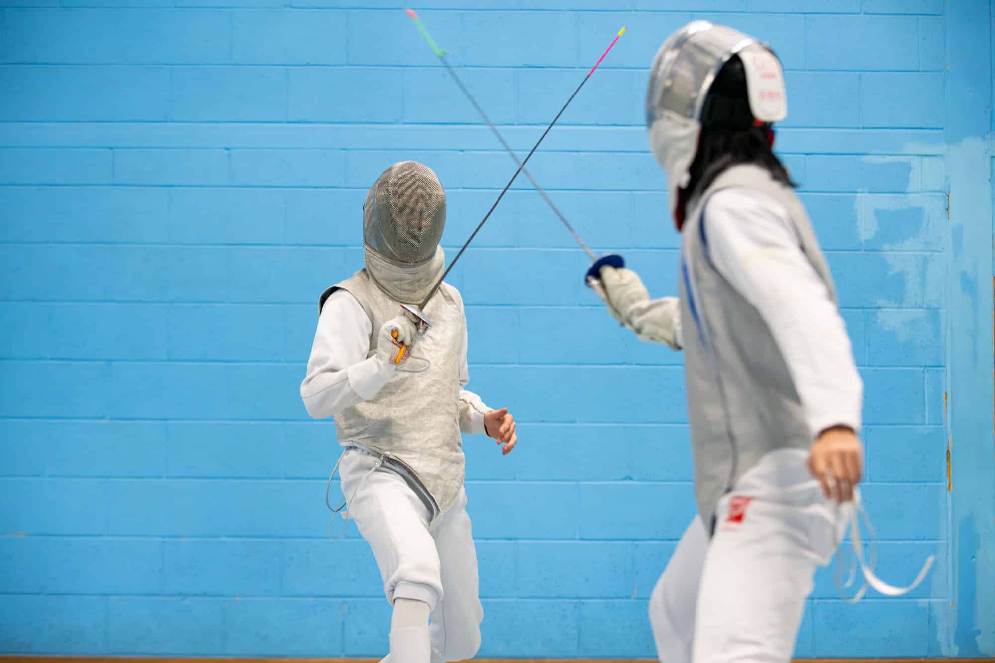 Two fencers in protective masks and white uniforms spar against a blue brick wall.