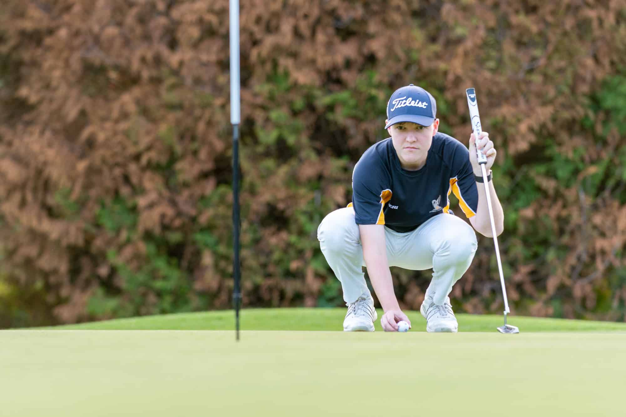 A golfer crouching on a green to read a putt, holding a putter in one hand and placing a ball with the other.