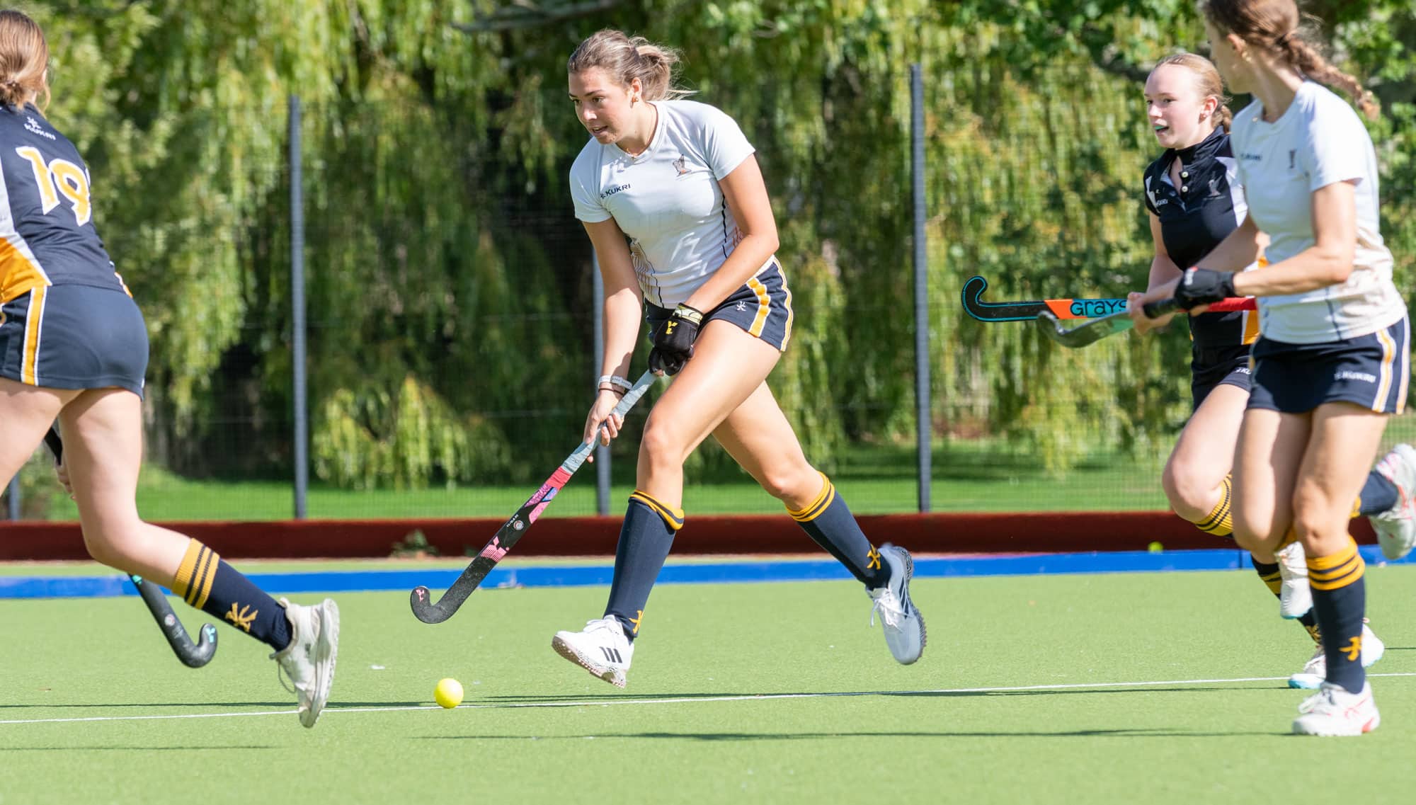 Female hockey players compete on a green turf pitch,
