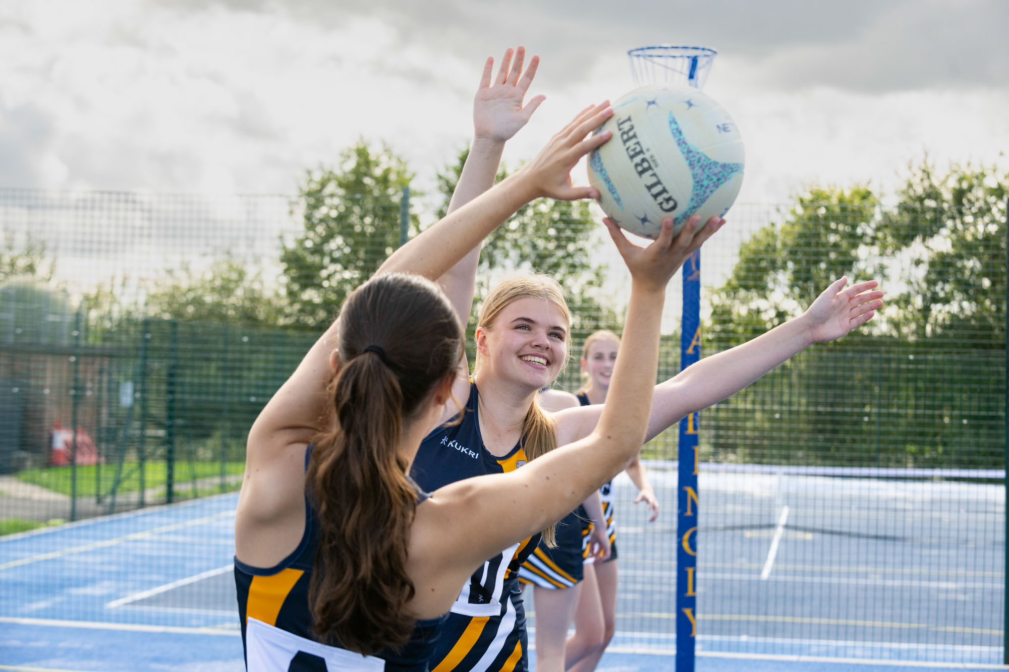 A netball player reaches up to catch the ball whilst being defended by an opponent, with two other players standing on the court in the background.