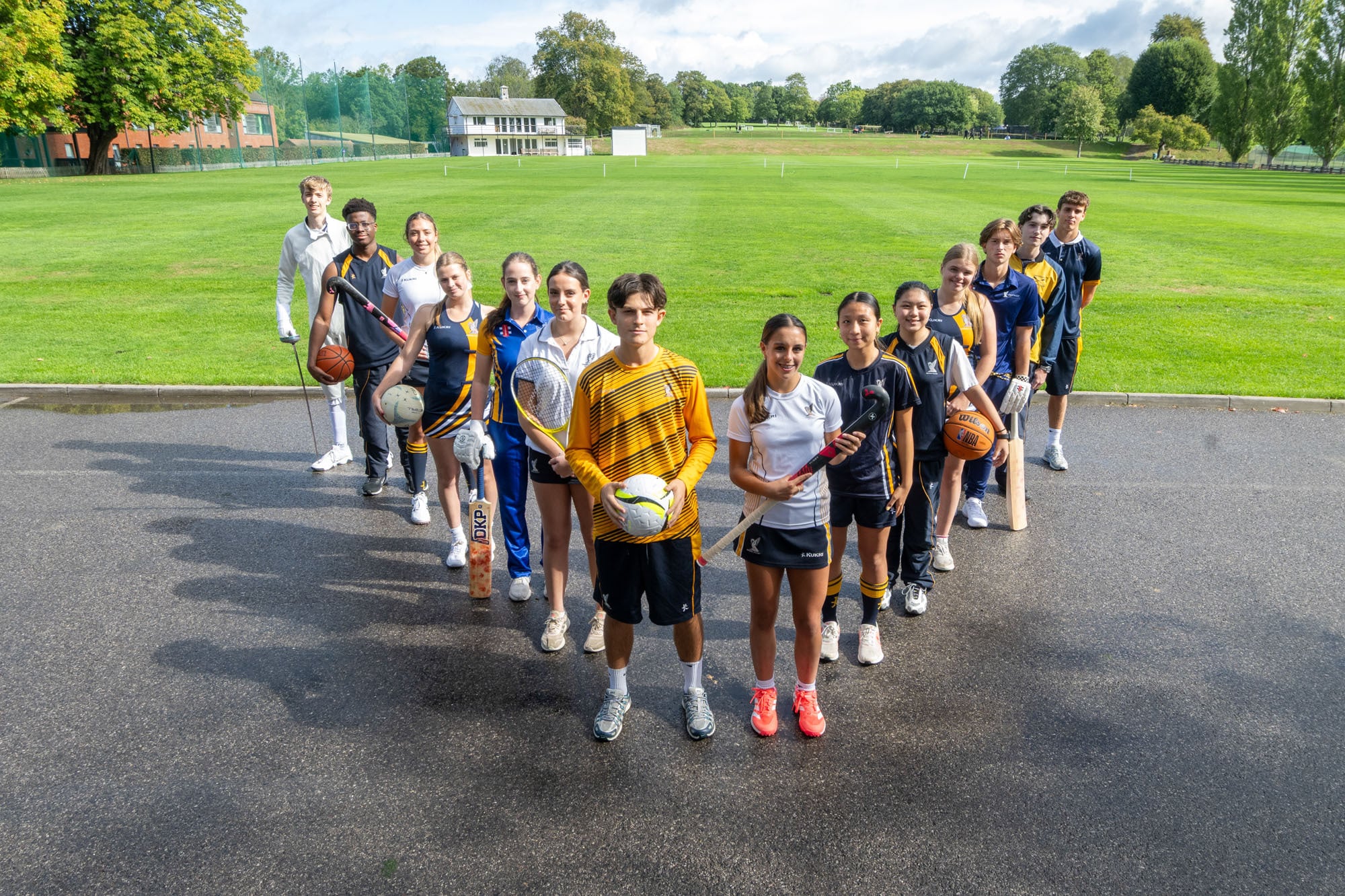 A diverse group of students posing on a paved court holding various sports equipment, including hockey sticks, cricket bats, and balls, with a large grassy field in the background.
