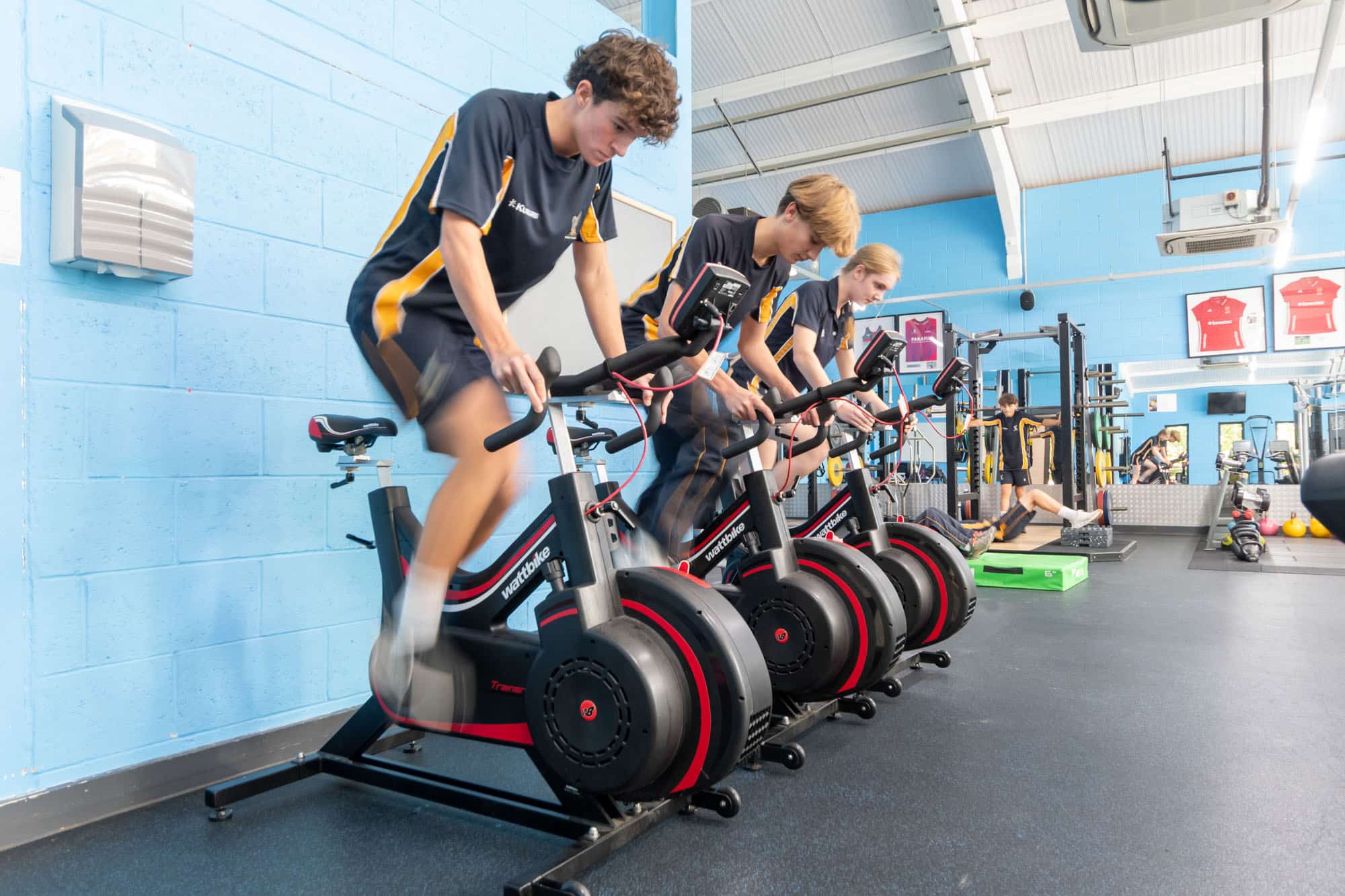 Three young athletes exercise on stationary bikes in a gym with blue walls.