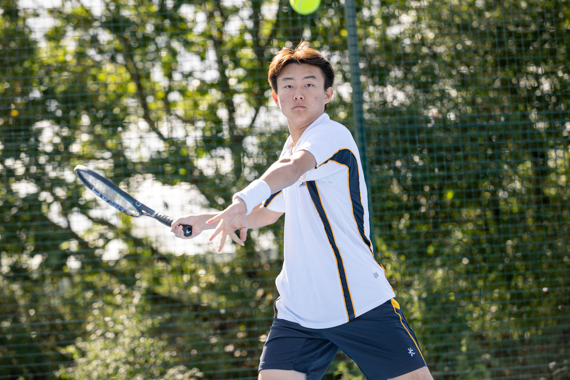 A female tennis player in a white polo shirt and skirt swings her racquet towards a yellow tennis ball on a court.