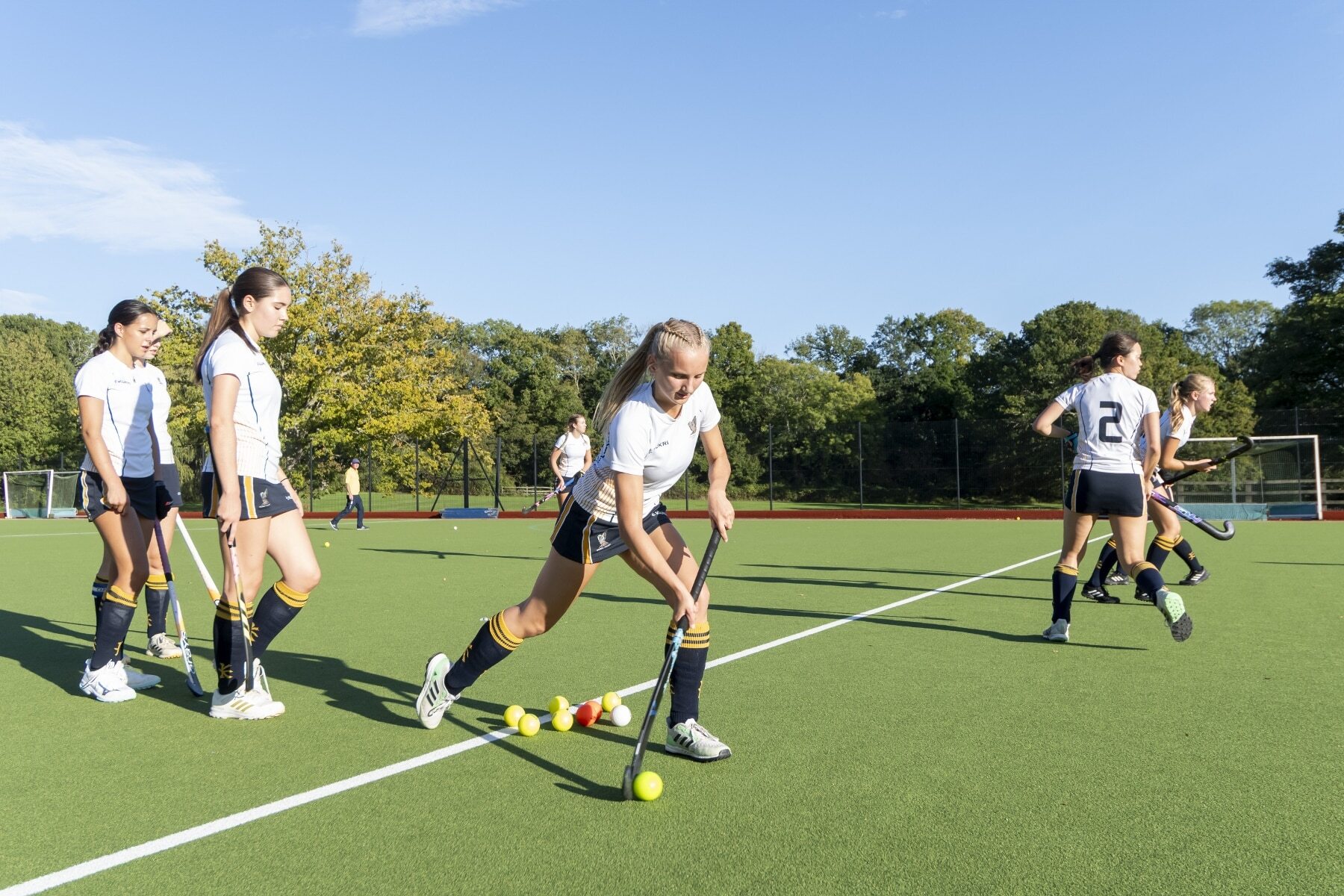 A field hockey player in a white kit dribbles a ball on a green pitch during practice with teammates nearby.