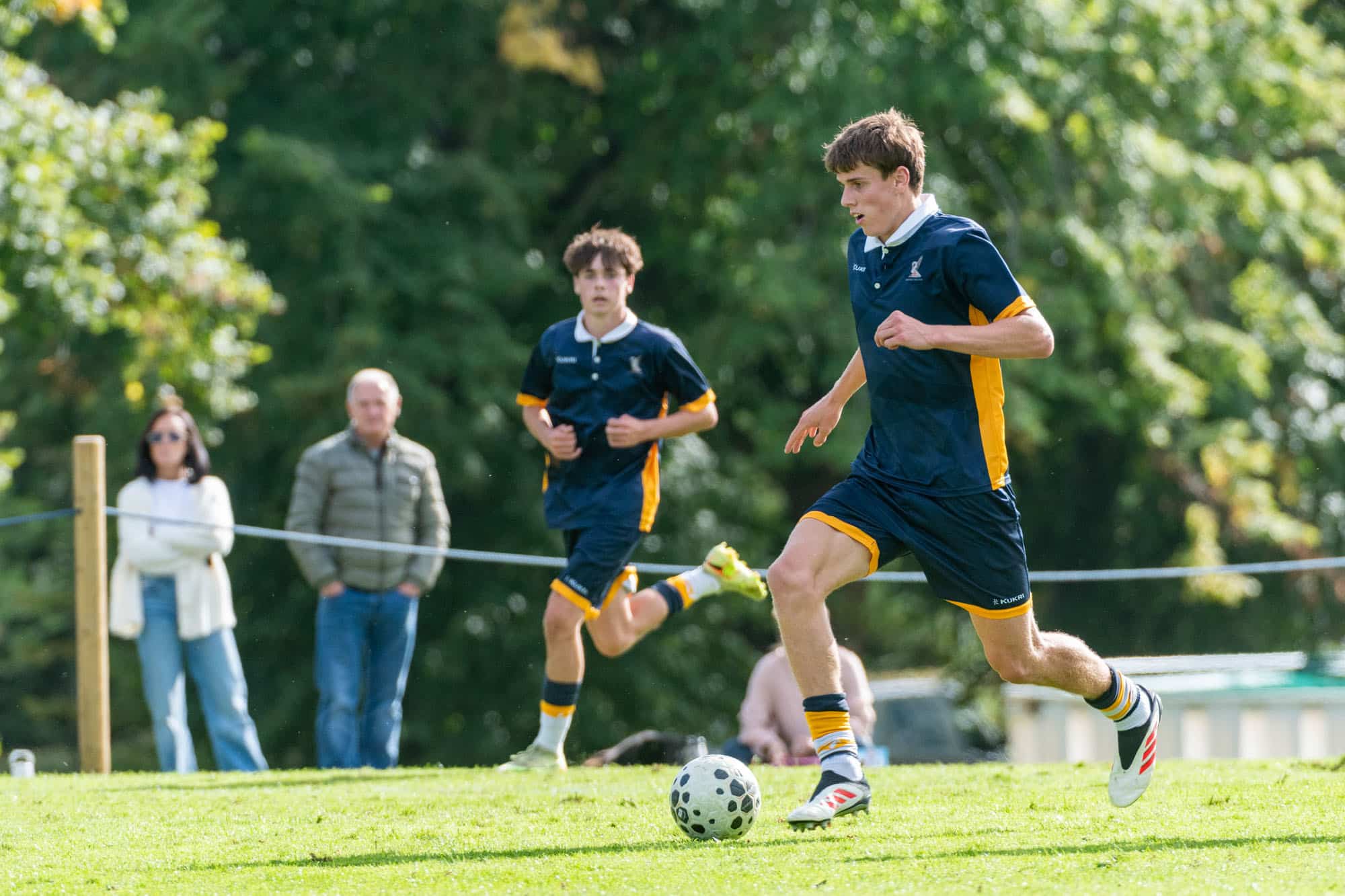 An Ardingly 1XI footballer in a blue and gold kit dribbles a ball across a grassy pitch whilst a teammate runs behind him.