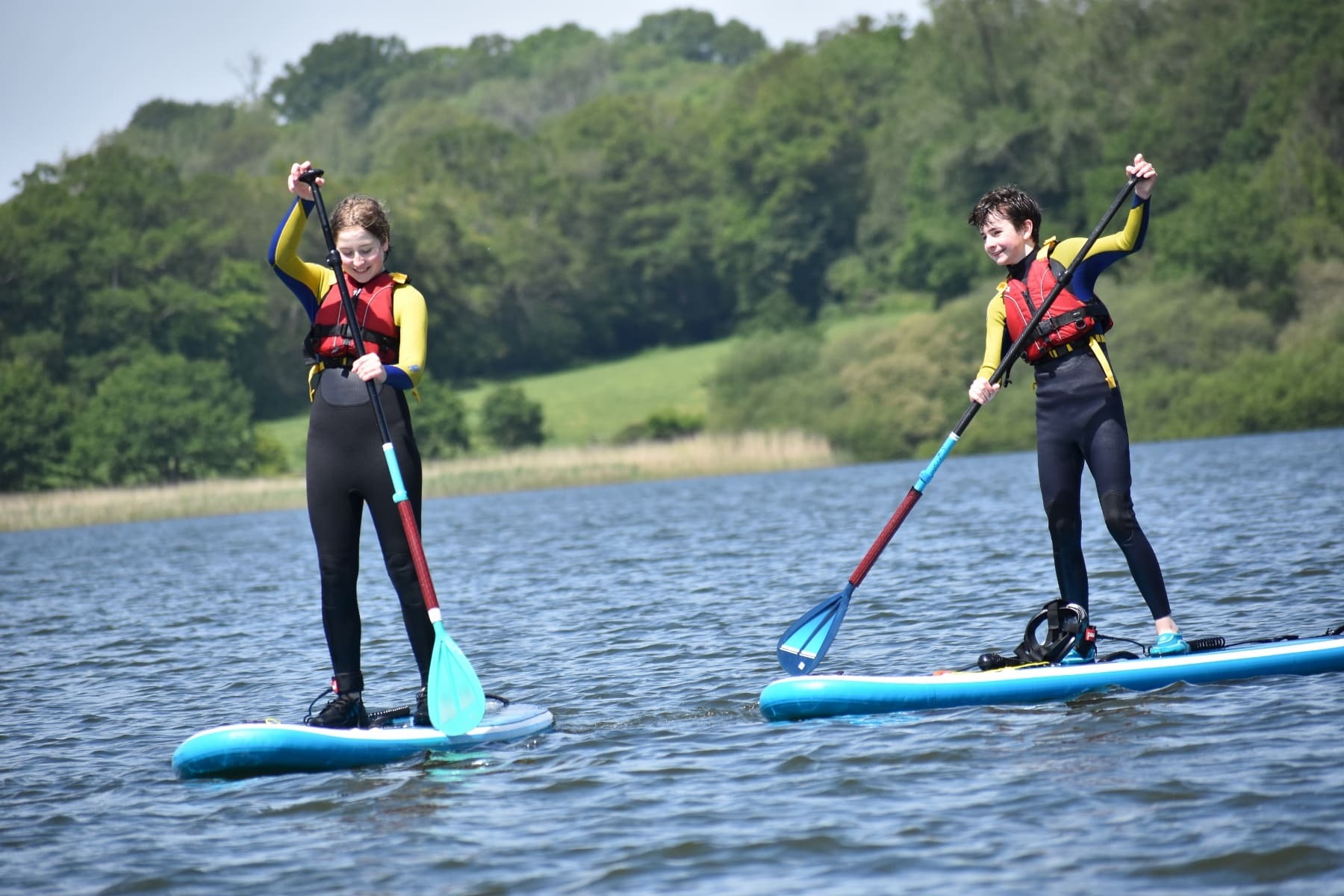 Two people in wetsuits and life jackets stand on paddleboards on a lake, both holding paddles.
